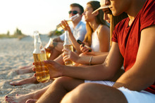 Group Of Friends Having Fun Enjoying Refreshing Beverage And Relaxing On The Beach At Sunset. Young Men And Women Drink Beer Sitting On A Sand In The Warm Summer Evening. Isolated View Of Bottles.
