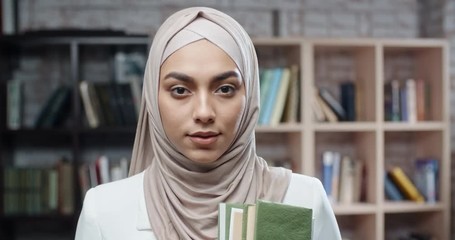 Wall Mural - Female student wearing hijab confidently looking at camera. spending time in campus library studying - modern muslim concept close up 4k