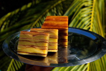 Bebinca, traditional Goan dessert from coconut milk dough, sweet Indian pudding on a metal plate, close up. Goan-Portuguese pie, delicious pieces in sunlight against palm leaves.