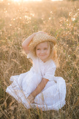 Cute little girl with blond long hair in a summer field at sunset with a white dress with a straw hat 