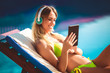 Portrait of young woman in the tropical sun near swimming pool on a deck chair using digital tablet