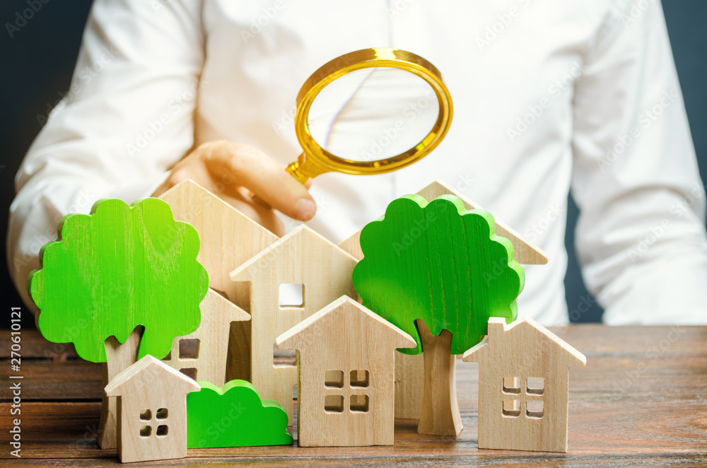 A man holds a magnifying glass over wooden houses and trees. Real estate valuation and selection of a place for the construction of new buildings. Evaluation of urbanism and residential space. - obrazy, fototapety, plakaty 