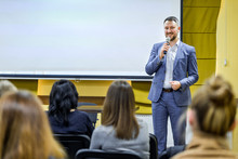 Business Leader Male Conducts A Lecture With A Smile On His Face During The Conference. Man In Stylish Blue Suit With Microphone Speaking On The Background Of White Screen.