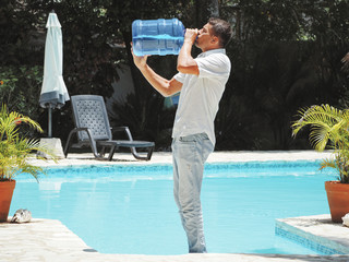 a young man wearing jeans and a shirt drinks water from a 20 liter bottle near the swimming pool. bright sun day is very hot.