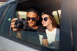 © artiemedvedev - Blogger in glasses filming his travel vlog on professional camera on road trip. Man photographer taking photos out of open window with girlfriend nearby. Multiracial hipsters couple on rear car seat.