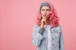 Young thinking nice woman with pink hair, stands over pink background copy space, looks up thoughtfully and touches the cheek with his finger, wears a white t-shirt and denim jacket.