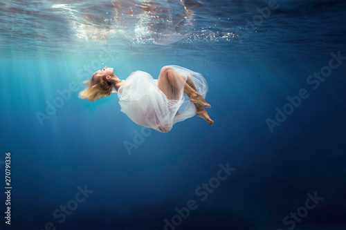 Fotomural Blonde girl wrapped in fine white cloth, sank in blue deep water of ocean, against dark sea background