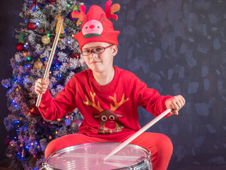 Happy Caucasian childish drummer in carnival costume Santa Claus Deer plays on a new drum set with drum sticks in his hands. Parents bought child drum as gift for Christmas. Lights on Christmas tree