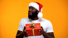 African-American Man In Santa Hat Showing Giftbox At Camera, Holiday Celebration