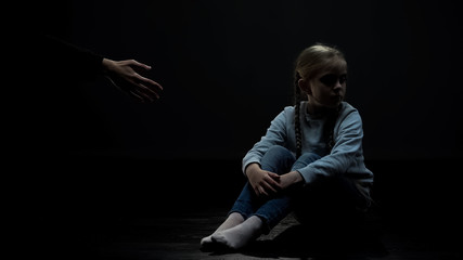 Wall Mural - Distrustful little girl sitting in dark room refusing adult helping hand, fear