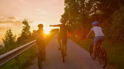 LENS FLARE: Group of cyclists rides ebikes along empty country road at sunset.