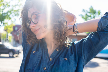 Outdoor close up portrait of young beautiful stylish happy smiling curly girl wearing sunglasses, posing in street. Sunny day light. Summer fashion concept. Copy, empty space for text