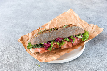 sandwich with baguette, ham, lettuce, kale on gray background, salad from kale and lettuce leaves and tomatoes on a white plate. takeaway sandwich, selective focus, copy space.