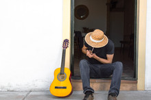 Young Man With Straw Hat And A Guitar Smoking A Marijuana Joint At The Door Of A White House In The Countryside. Guitarist Sitting In The Street.