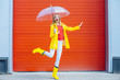 © Iryna - Emotional woman dancing laughing under umbrella in yellow raincoat, rubber boots, red t-shirt over red garage door background. Cool expression on face. Outdoor. Full height. Weather forecast concept
