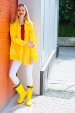 Full Length Portrait Of A Smiling Blond Teenager Woman Dressed In Yellow Raincoat And Rubber Boots Posing While Standing On Parking Garage Red Door On Background. Outdoor Lifestyle Concept