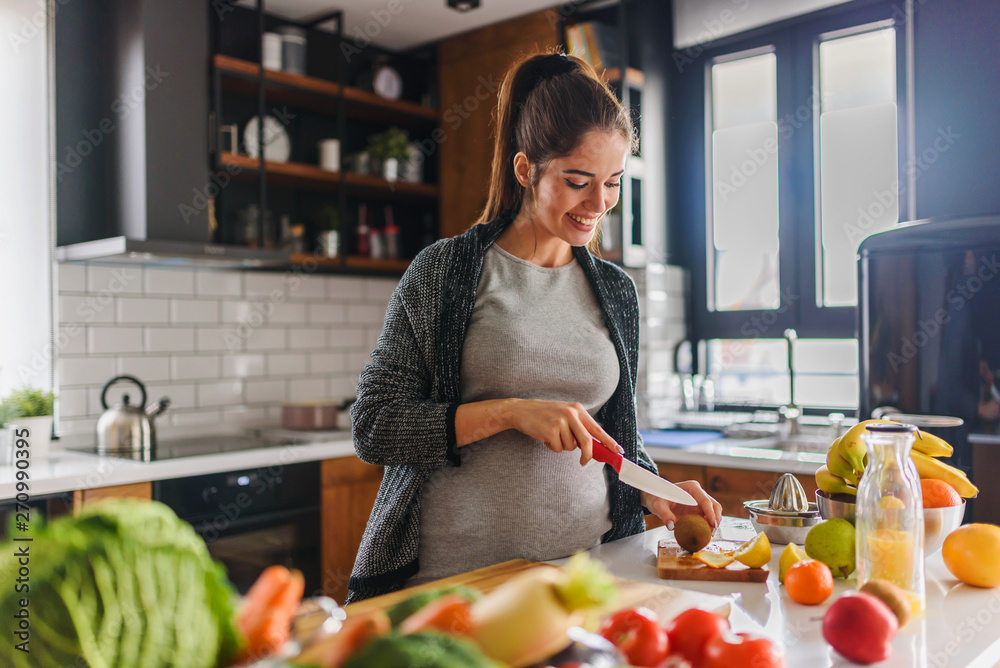 Beautiful smiling young pregnant woman preparing healthy food with lots of fruit and vegetables at home kitchen - obrazy, fototapety, plakaty 