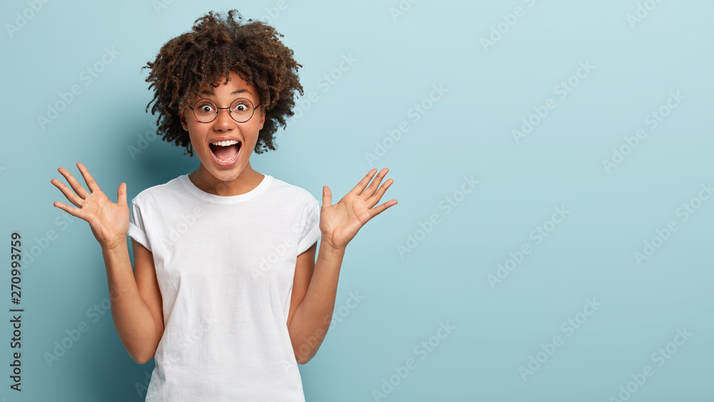 Curly optimistic woman raises palms from joy, happy to receive awesome present from someone, shouts loudly, dressed in casual white t shirt, isolated on blue background. Excited Afro female yells - obrazy, fototapety, plakaty 