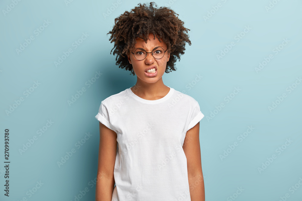 Pissed annoyed woman has Afro hairstyle, clenches teeth from anger, stares with hate, frowns face, wears round specacles and white casual t shirt, models on blue background, expresses hateful emotions - obrazy, fototapety, plakaty 