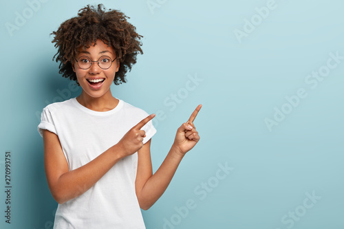 Fotografía Happy smiling woman seller points at upper right corner, advertises item on blank space, has friendly facial expression, wears casual white t shirt, isolated on blue background