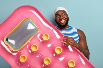 Mirthful happy Afro man with bright toothy smile, holds inflatable mattress, ready to relax during sunny day at beach, wears white swimcap, has fun during summer vacation, enjoys pool resort