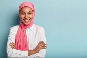 Wall Mural - Studio shot of cheerful religious Muslim woman keeps arms folded, smiles broadly, has white teeth, expresses positive emotions, satisfied after praying, models over blue background with blank space