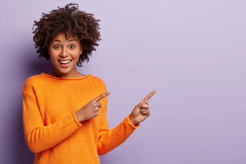 Photo of delighted African American woman points away with both index fingers, promots awesome place for your advertising content, isolated over purple background, gives advice, shows copy space