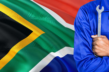 Mechanic In Blue Uniform Is Holding Wrench Against Waving South Africa Flag Background. Crossed Arms Technician.