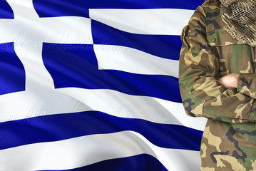 Crossed arms Greek soldier with national waving flag on background - Greece Military theme.