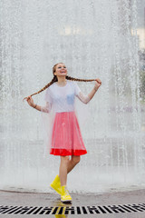 Canvas Print - Young wet pretty girl with two braids in yellow boots and with transparent umbrella stands inside of fountain.