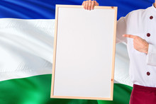 Chef Holding Blank Whiteboard Menu On Lesotho Flag Background. Cook Wearing Uniform Pointing Space For Text.