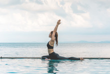 Happy Asian Woman Wearing Black Sport Wear Practice Yoga Pigeon Pose On The Pool Above The Beach With Beautiful Sea In Tropical Island,Feeling Comfortable And Relax In Holiday,Vacations Concept