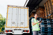 © twinsterphoto - Young Asian male logistics warehouse distribution business entrepreneur wearing reflective jacket using a digital tablet. He is surrounded by plenty of pallets and truck in shipping cargo.