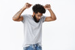 © Cookie Studio - Handsome and cool african american adult guy with curly hair, beard tattoos and pierced nose looking down as dancing with raised hands having fun catching rhythm of music over gray wall