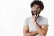 © Cookie Studio - African american guy in glasses making plan in mind thinking up grocery list holding hand on beard looking left thoughtful, frowning as considering what choice right creating new idea over gray wall