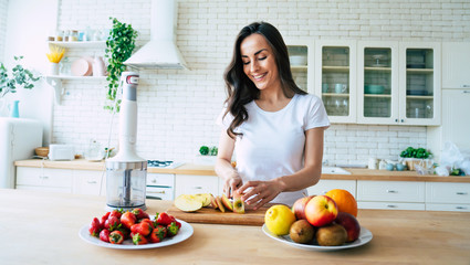 Beautiful woman making fruits smoothies with blender. Healthy eating lifestyle concept portrait of beautiful young woman preparing drink with bananas, strawberry and kiwi at home in kitchen.