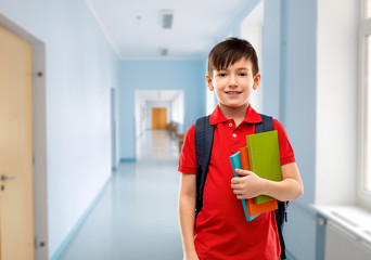 Wall Mural - education and people concept - smiling little student boy in red polo t-shirt in glasses with books and bag over school corridor background