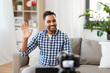 © Syda Productions - blogging, videoblog and people concept - indian male blogger with camera recording video and waving hand at home