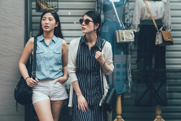 Wall Mural - two beautiful young asian chinese women in city shopping street trendy wear looking away leaning on shop window with dummy inside. female tourists lifestyle exterior with clothing store find search