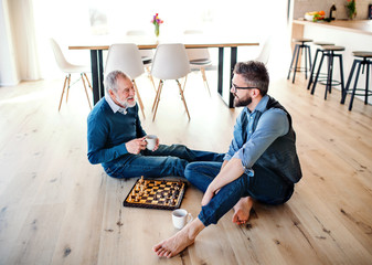 Wall Mural - An adult hipster son and senior father sitting on floor indoors at home, playing chess.