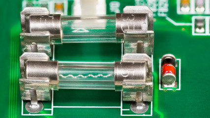 Miniature glass electric fuses detail with wavy fusible wire. Metal transparent current protector, red diode and blurry electronic components soldered on green circuit board. Electrical safety device.