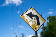 Old, Dilapidated Yellow Curve Ahead Road Sign With Peeling Paint Against Blue Sky With Power Lines
