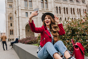 Wall Mural - Blissful white female model in retro jeans making selfie with smile. Lovable caucasian girl in hat expressing positive emotions while taking picture of herself in old part of city.