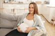 Family, maternity and childbirth concept. Picture of adorable young brunette pregnant woman in casual wear sitting comfortably on geay couch in living room, holding her large belly, smiling