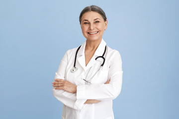 Isolated studio image of charismatic beautiful Caucasian senior female chief doctor crossing arms on her chest confidently and smiling broadly dressed in white overall with stethoscope around her neck