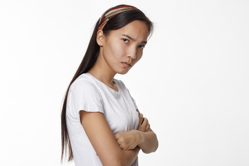 Grumpy dissatisfied young Chinese woman in white top frowning and crossing arms on her chest, being offended by her friends, expressing negative emotions, posing in studio in closed posture