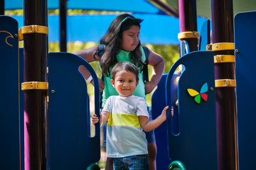 Young boy 3 years of age is standing and holding on to playground slide with older sister standing behind to help.