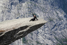Fearless Woman Sitting On Trolltunga Rock Formation And Waving Hand. Jutting Cliff Is In Odda, Hordaland County, Norway