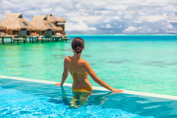 Wall Mural - Luxury overwater bungalow hotel room with infinity swimming pool woman looking at blue ocean view. Tropical travel summer vacation lifestyle.