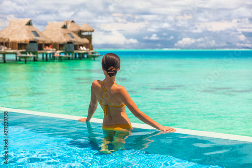 Luxury overwater bungalow hotel room with infinity swimming pool woman looking at blue ocean view. Tropical travel summer vacation lifestyle.